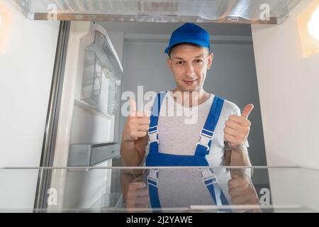 Un homme souriant en salopette montre ses pouces. Concept de réparation de réfrigérateur. Photo de l'intérieur du réfrigérateur Banque D'Images