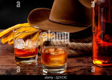 Le thème du concept de l'Ouest sauvage avec chapeau de cowboy, corde lasso, gants en cuir, deux verres de whisky sur les rochers et bouteille de bourbon sur table en bois Banque D'Images