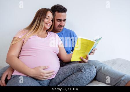 jeune couple hétérosexuel latin caucasien en pyjama assis sur le lit lisant un livre, homme barbu et femme enceinte dans la chambre lisant ensemble. Banque D'Images