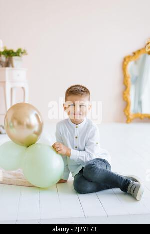 Un enfant garçon est assis sur le sol et tient des ballons dans sa main à la maison et souriant Banque D'Images