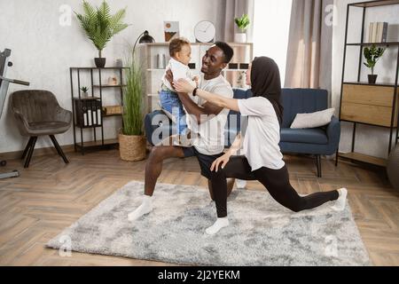 Jeune heureux couple multiracial faisant du yoga sur la quarantaine à la maison. Un jeune homme afro-américain et sa femme musulmane debout dans un asana alors qu'un homme tient leur petit fils dans ses bras. Banque D'Images