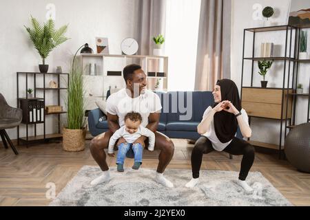 L'Homme africain athlétique musculaire tient son fils sur ses épaules et belle condition physique la femme musulmane dans les vêtements d'entraînement font des exercices de squat dans leur appartement lumineux et spacieux. Banque D'Images