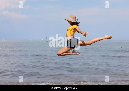 jeune fille avec chapeau de paille effectue un saut très élevé de gymnastique rythmique sur le bord de mer Banque D'Images