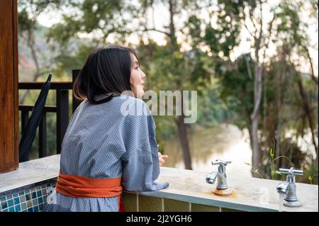 Bonne jeune femme asiatique en peignoir assis sur la piscine naturelle de source chaude au milieu de la nature à la station Banque D'Images