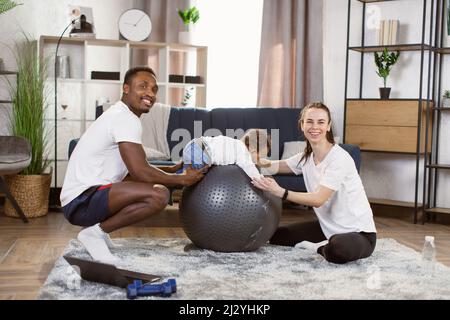 Âge moyen beau caucasien sportswoman avec l'homme afro-américain et leur race mixte mignon fils faisant l'exercice de yoga en utilisant le ballon de fitness à la maison. Banque D'Images