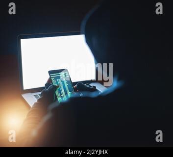 Accès à tous vos mots de passe. Photo d'un pirate méconnaissable à l'aide d'un téléphone portable et d'un ordinateur portable dans l'obscurité. Banque D'Images