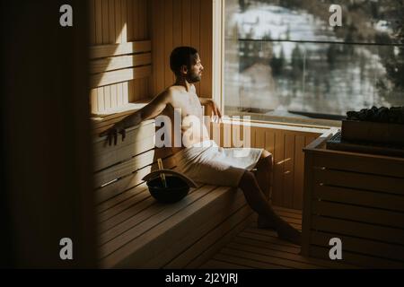 Beau jeune homme se détendant dans le sauna et regardant la forêt d'hiver par la fenêtre Banque D'Images