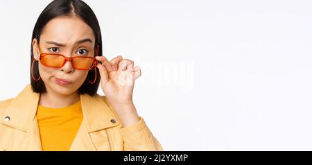 Image de jeune femme asiatique élégante, regardant sous des lunettes de soleil avec le visage sceptique, incrédulité, exprimer le doute, debout sur fond blanc Banque D'Images