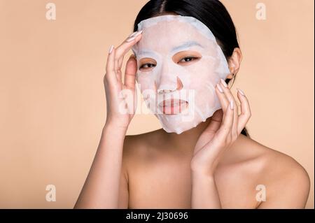 Procédures de soin du visage. Gros plan de la jeune femme asiatique appliquant un masque hydratant facial en coton sur le visage, prend soin de la peau, prévient les rides, regarde l'appareil photo, se tient sur un arrière-plan beige isolé Banque D'Images