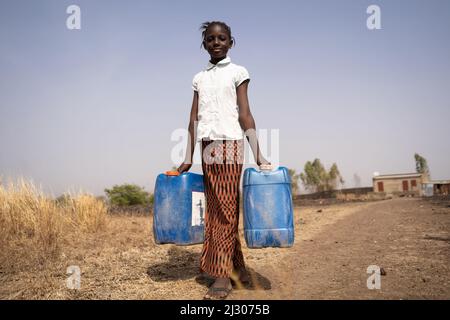 Petite fille africaine skinny transportant deux conteneurs d'eau au milieu d'un paysage aride; concept de pauvreté, manque de raccords de tuyaux dans privé rural ho Banque D'Images