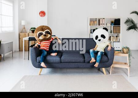 Deux enfants portant un masque géant pour animaux assis sur le canapé. Banque D'Images