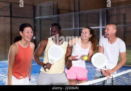 Rire des hommes et des femmes avec des raquettes et des balles qui parlent sur un terrain de padel intérieur Banque D'Images