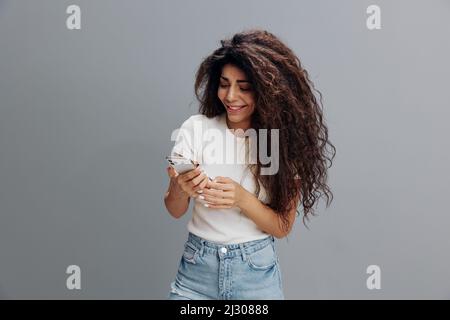 Appareils modernes et communication en ligne. Bonne jeune fille latine à cheveux afro-cheveux en t-shirt blanc avec cheveux bouclés regardant dans le smartphone et blogging Banque D'Images