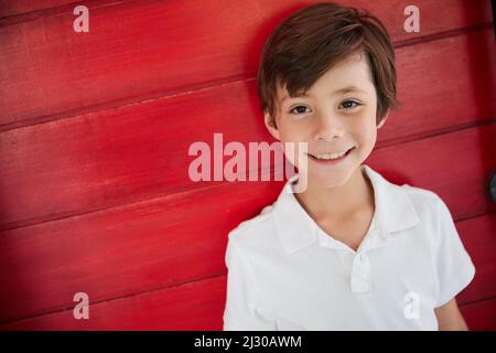 HES un adorable petit gars. Portrait d'un petit garçon souriant debout contre un mur rouge. Banque D'Images