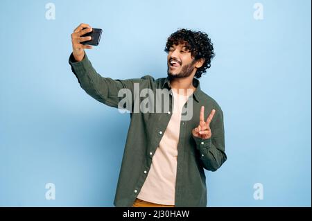 Joyeux Indien positif ou arabe gars excité, dans une tenue décontractée et élégante, faisant selfie tourné sur le téléphone portable et souriant dans l'appareil photo, montre le symbole de paix, posant sur un arrière-plan bleu isolé Banque D'Images