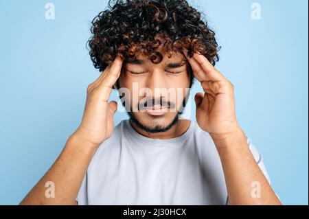 Gros plan d'un homme indien malsain triste avec les yeux fermés, massant ses temples, éprouvant une migraine, souffrant de maux de tête, surtravaillé, en cas de besoin de repos ou de traitement, tenez-vous isolé sur fond bleu Banque D'Images