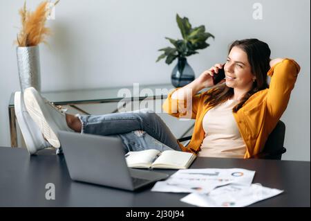 Positive réussie Femme de caucasien, employé de bureau, directeur de création, assis dans une chaise dans une posture détendue avec ses pieds sur la table, parlant un téléphone mobile pendant une pause de travail, regarde loin, sourit Banque D'Images