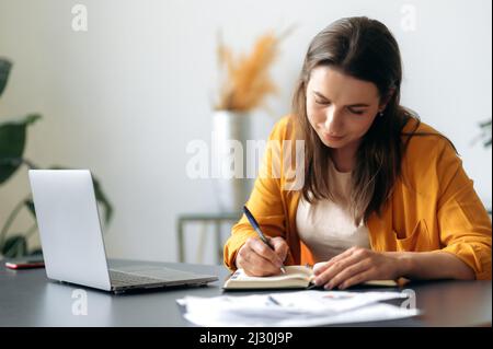 Focalisé réussi SMART et déterminé jeune femme d'affaires caucasienne, responsable, étudiant ou indépendant, assis à la table, portant des vêtements élégants, utilisant un ordinateur portable, prendre des notes, analyser, e-learning Banque D'Images