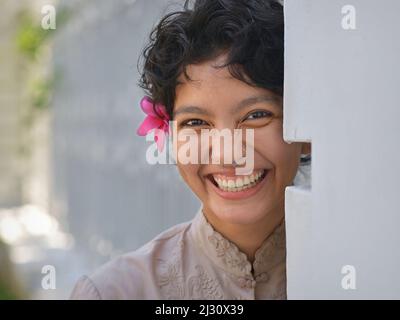 Belle belle jeune femme gaie Latina avec de courts cheveux noirs bouclés et une fleur rouge dans ses cheveux sourit au spectateur autour d'un coin. Banque D'Images