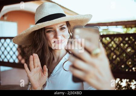 jeune femme souriante en chemise blanche avec chapeau ayant une réunion vidéo sur un smartphone dans le patio de l'hôtel maison d'hôtes. Banque D'Images