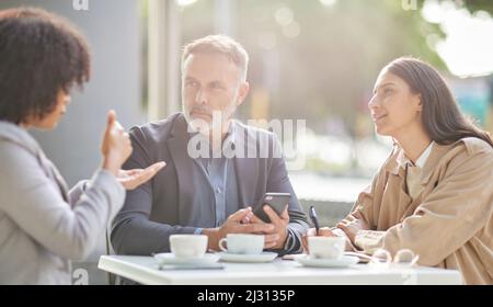 Je suis heureux que vous ayez soulevé cette question. Photo d'un groupe de collègues qui remue des idées dans un café tout en utilisant leur smartphone. Banque D'Images