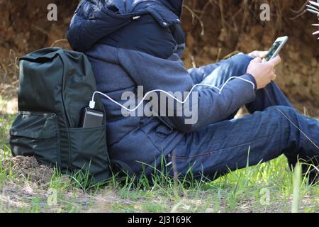 Un homme assis au sol, tient un smartphone dans ses mains et le charge avec une banque d'énergie noire sur fond de vue sur la nature. Appareil portable de voyage Banque D'Images