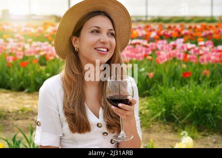 Jeune belle femme avec chapeau de paille boire un verre de vin rouge sur fond fleuri regardant à côté avec visage heureux souriant Banque D'Images