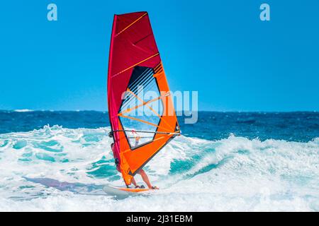 sports d'été: planche à voile sur les vagues pendant un après-midi ensoleillé d'été avec la voile rouge et orange sur l'eau bleue de l'océan. Banque D'Images