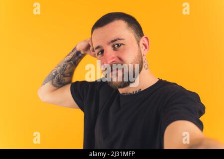 Homme européen attrayant avec une moustache à cheveux noirs courts barbe et des tatouages portant un T-shirt noir et prenant un selfie jaune fond studio prise de vue moyenne gros plan. Photo de haute qualité Banque D'Images