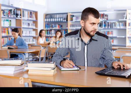 Un gars concentré se préparant à l'examen dans la bibliothèque de l'université, lisant le livre et utilisant l'ordinateur portable pour la recherche d'informations Banque D'Images
