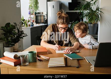 Jeune mère montrant comment écrire à son fils pendant qu'ils font leurs devoirs ensemble à table dans la cuisine Banque D'Images