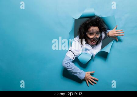 Scientifique humoristique stupide avec expression de bande dessinée sur fond bleu. Jeune adulte chimiste avec un look wacky, cheveux désordonnés et sale visage agissant goofy après l'explosion d'expérience. Prise de vue en studio. Banque D'Images