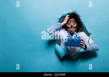 Scientifique fou au visage sale et à l'expression de goofy ayant stand rempli d'échantillons. Un chimiste fou avec un aspect amusant et des cheveux enrasés qui collent la langue tout en ayant un rack rempli de tubes à essai en plastique. Banque D'Images