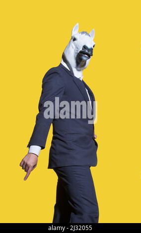 Photo verticale d'un homme créatif en costume avec un masque blanc posé sur un mur de fond jaune. Banque D'Images