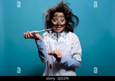 Un ouvrier de laboratoire insase grinçant la crème tout en égouttant du composé toxique de la pipette dans un tube à essai en verre tout en regardant l'appareil photo sur fond bleu. Scientifique fou avec des cheveux salissants et visage sale en utilisant la pipette Banque D'Images