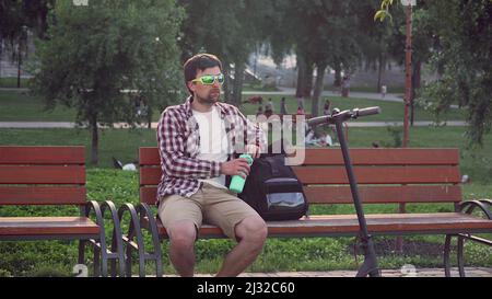 L'homme caucasien est assis sur un banc de parc après avoir porté un scooter électrique et boire d'une bouteille turquoise tout en l'emportant de son sac à dos. Homme Banque D'Images