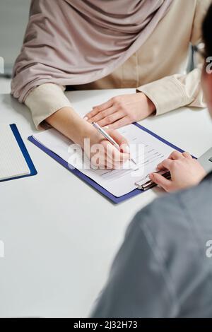 Main d'une jeune patiente musulmane avec un stylo pointant vers le formulaire médical sur le presse-papiers tout en remplissant les informations personnelles dans les cliniques Banque D'Images