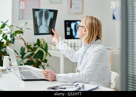 Femme blonde radiologue en blanchon regardant l'image radiographique des poumons du patient tout en étant assise devant l'ordinateur portable pendant la consultation en ligne Banque D'Images