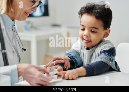 Pédiatre contemporain mettant l'oxymètre de pouls sur le bout du doigt de mignon petit patient pour vérifier le niveau de saturation en oxygène dans son sang Banque D'Images