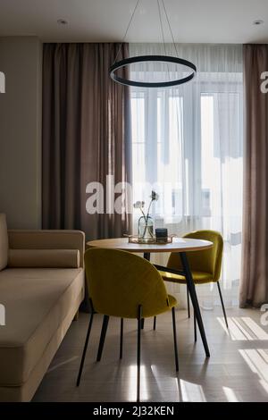 Deux chaises jaune moutarde, debout près d'une table ronde, près d'un canapé beige et de rideaux blancs et gris sur la fenêtre, dans une cuisine spacieuse Banque D'Images