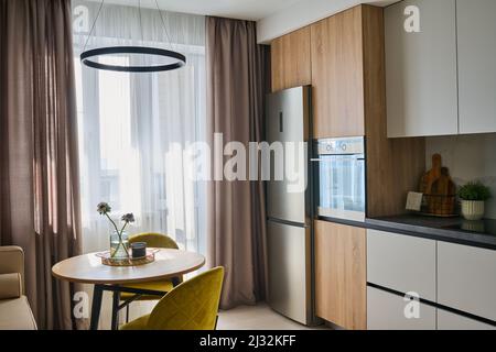 Table de cuisine ronde et deux chaises confortables de couleur jaune moutarde entre les fenêtres avec rideaux pastel et réfrigérateur Banque D'Images