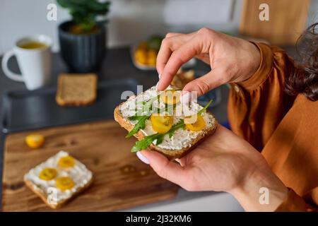 Mains de jeunes végétariennes mettant des tomates cerises jaunes hachées et des feuilles de ruccola sur le sandwich pour le petit déjeuner Banque D'Images