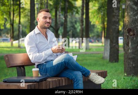 Un homme souriant est assis sur un banc dans le parc, compte des dollars. Un jeune homme sur fond d'arbres verts, une chaude journée d'été ensoleillée. Lumière douce et chaude, gros plan. Banque D'Images