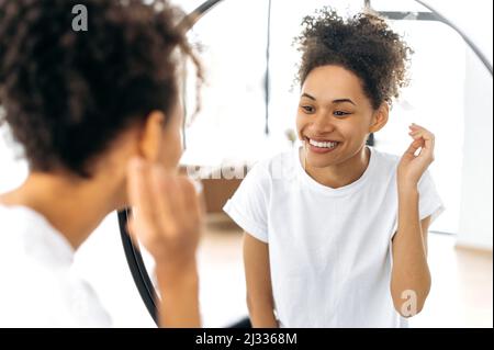 Joyeuse belle fille afro-américaine heureuse dans un t-shirt blanc de base, se tient devant un miroir le matin, s'admire elle-même, sourit, commence la journée bien, bonne humeur Banque D'Images