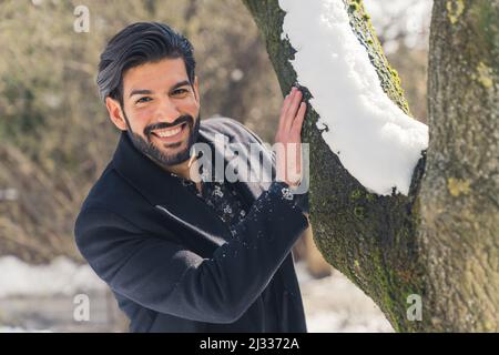 Élégant et beau Latin Man en manteau et costume noir jouant cacher et chercher derrière l'arbre dans un parc pendant les vacances d'hiver. Photo de haute qualité Banque D'Images