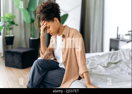 Bouleversée afro-américaine jeune fille, élégamment habillée, assis sur le lit à la maison, préoccupé par les problèmes personnels, triste par les mauvaises nouvelles, éprouvant la dépression, le désespoir, les maux de tête, a besoin de repos et de soutien psychologique Banque D'Images