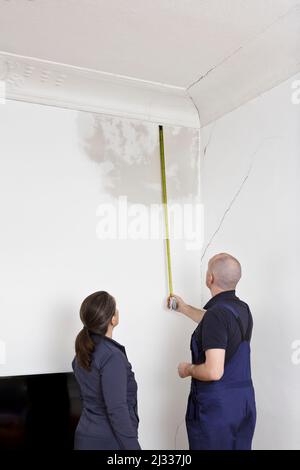 Concierge ou gardien de bâtiment mesurant l'étendue d'une grande tache d'eau sur le mur fissuré d'un ancien bâtiment, observation des locataires féminins. Banque D'Images