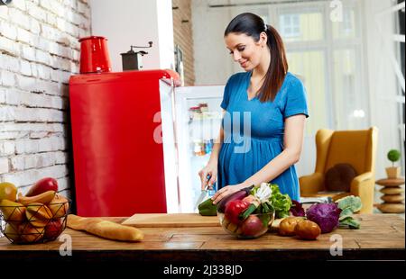 Bonne femme enceinte à la maison dans la cuisine avec salon en arrière-plan préparant la salade, manger des aliments sains à partir de légumes. Banque D'Images