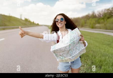 Jolie jeune femme avec sac à dos et carte hitchranking sur la route, montrant le pouce vers le haut, la vérification de l'itinéraire de voyage, l'espace libre Banque D'Images