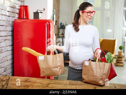 Femme enceinte dans la cuisine déballant des sacs d'épicerie pleins de légumes arrivant à la maison de shopping. Banque D'Images